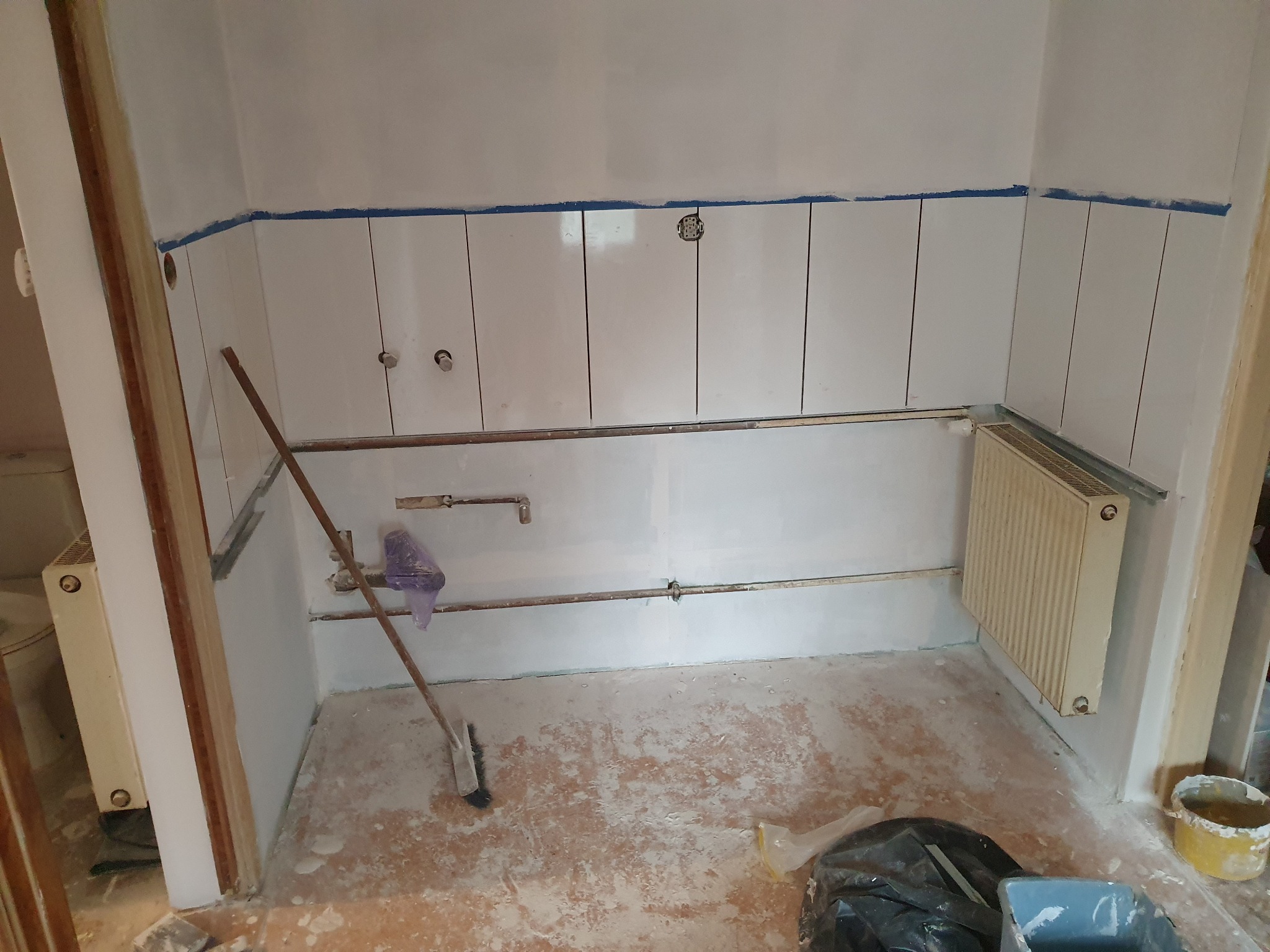 Wnętrze w trakcie remontu: ściany z częściowo położonymi białymi płytkami zabezpieczonymi niebieską taśmą, widoczne rury, grzejnik, oraz narzędzia i materiały budowlane na podłodze.