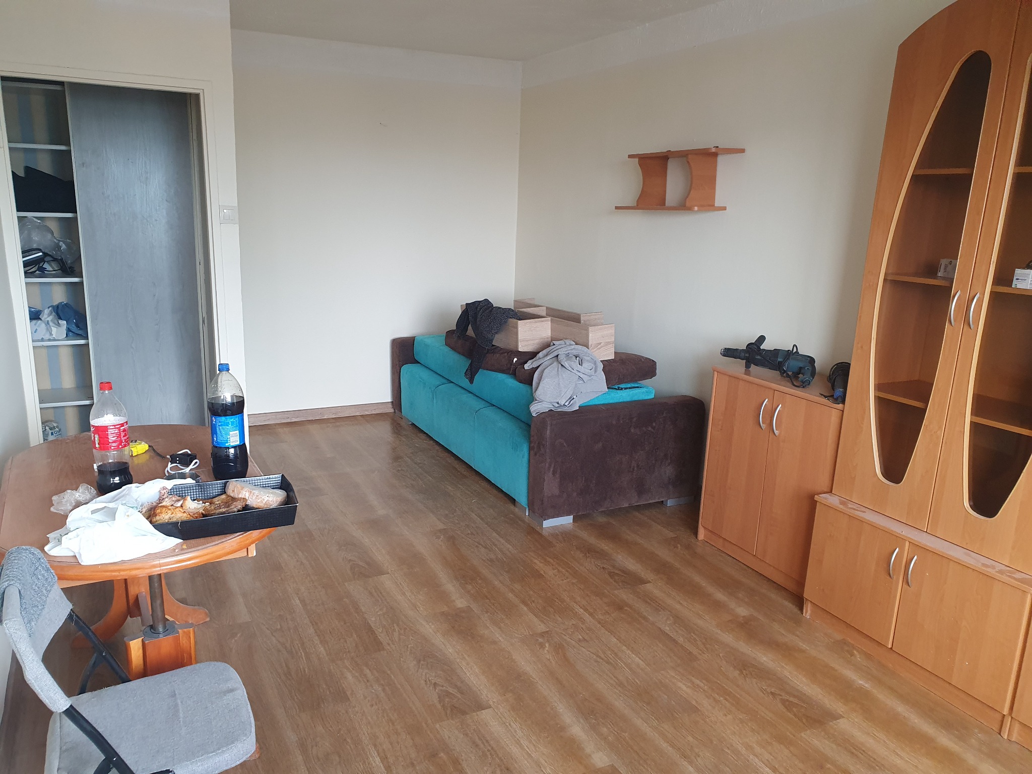 Wnętrze pokoju z drewnianą podłogą, turkusową kanapą przy ścianie, drewnianą szafką i stołem z jedzeniem, w tle białe ściany i drzwi do garderoby.