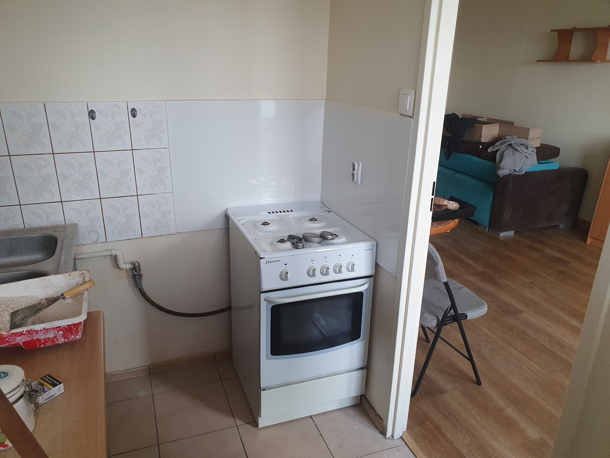 Wnętrze kuchni w trakcie remontu, widoczny biały piec, fragment zlewu, puszka z farbą i narzędzia malarskie na blacie, płytki ścienne częściowo zakryte białą farbą, w tle widok na pokój z meblami...