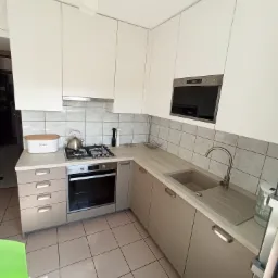 Nowoczesna kuchnia z białymi szafkami górnymi i beżowymi dolnymi, z wbudowanym piekarnikiem i mikrofalówką, oraz szarym zlewozmywakiem i baterią.