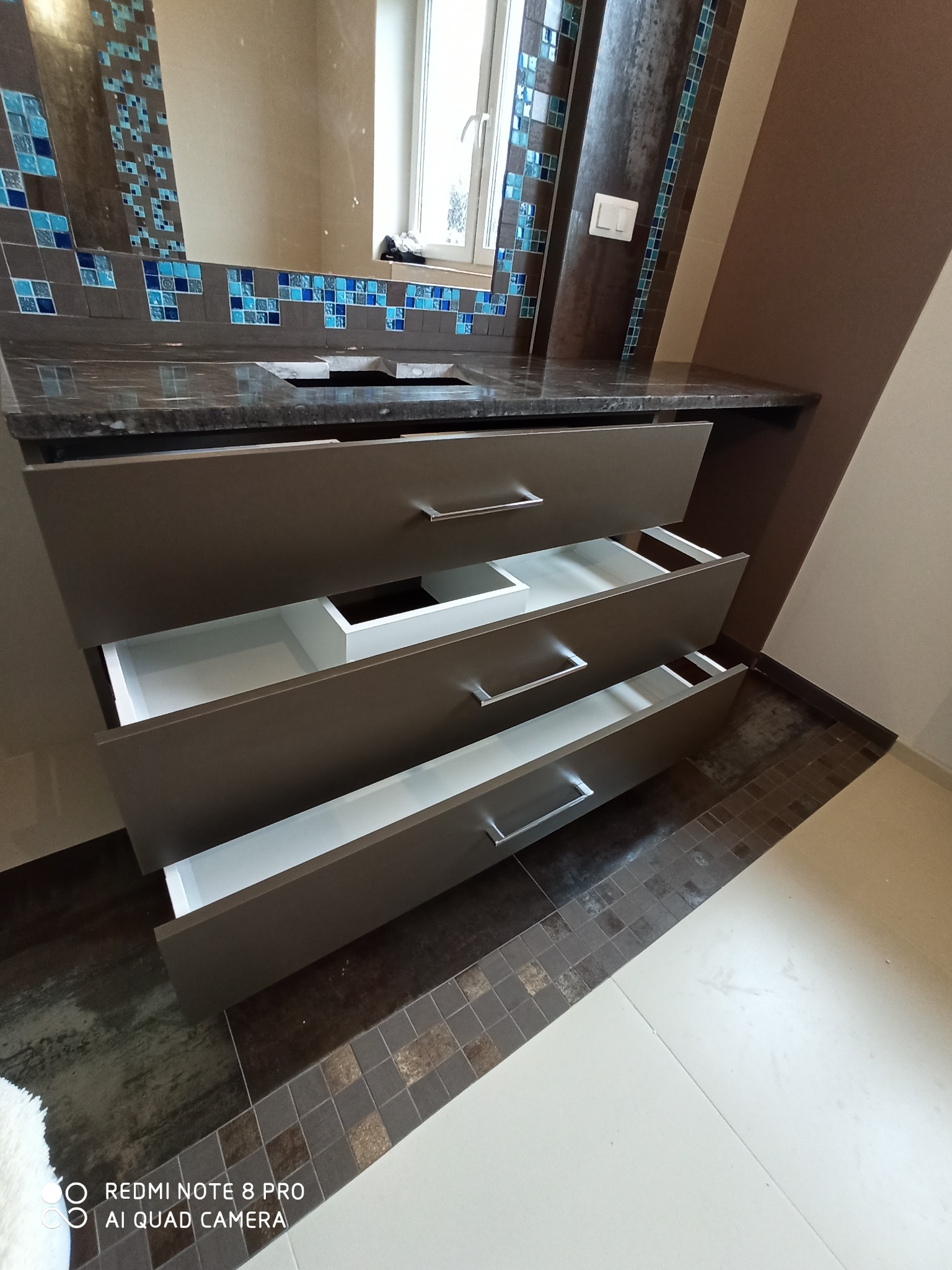 Szafka łazienkowa na wymiar z trzema wysuniętymi szufladami, szare fronty z poziomymi uchwytami, blat z ciemnego granitu, mozaika z niebieskich płytek nad blatem.