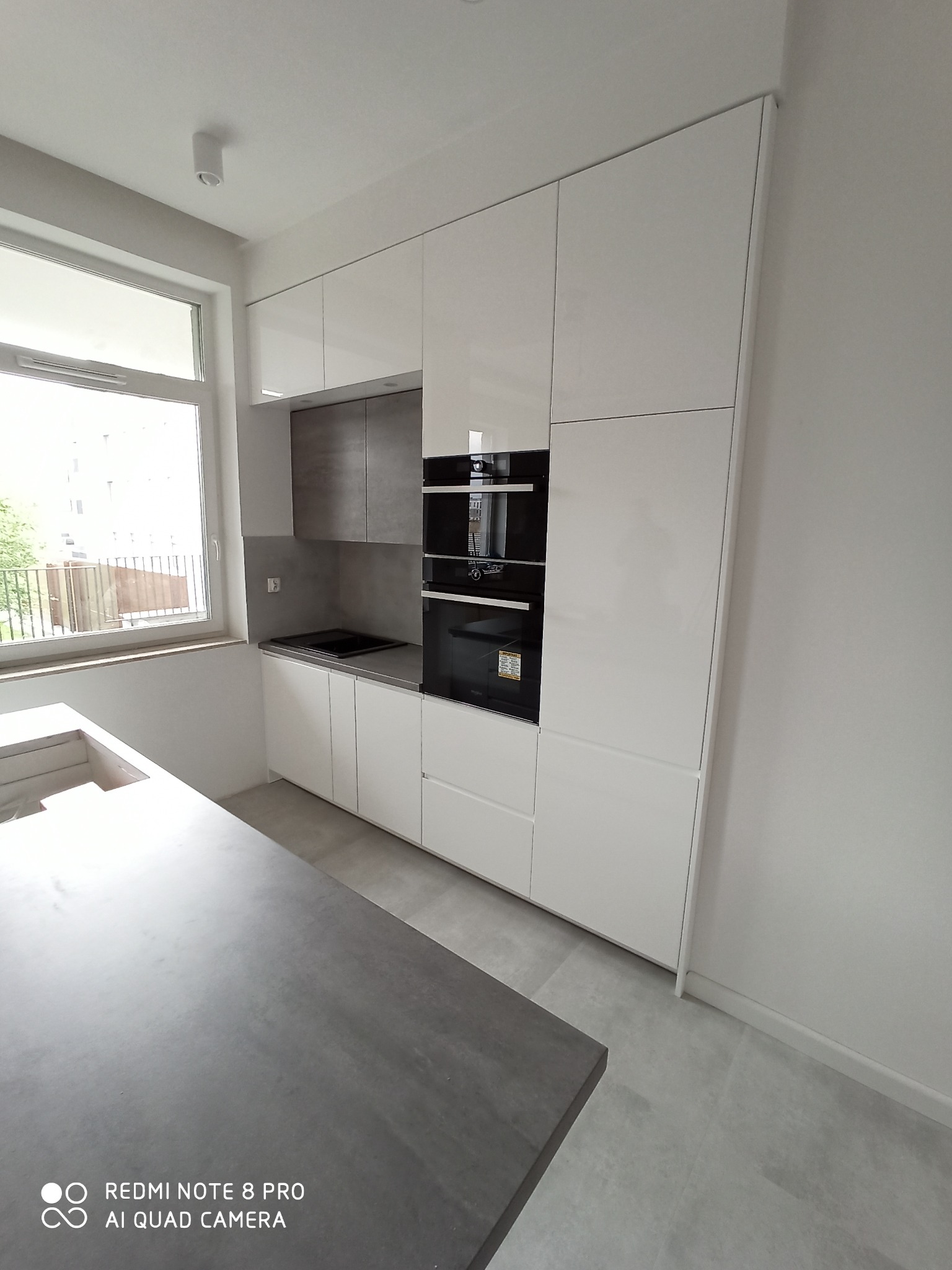 Nowoczesna kuchnia z białymi, lakierowanymi szafkami i szarymi elementami nad blatem roboczym, z wbudowanym piekarnikiem i mikrofalówką, widok na wyspę kuchenną i okno.