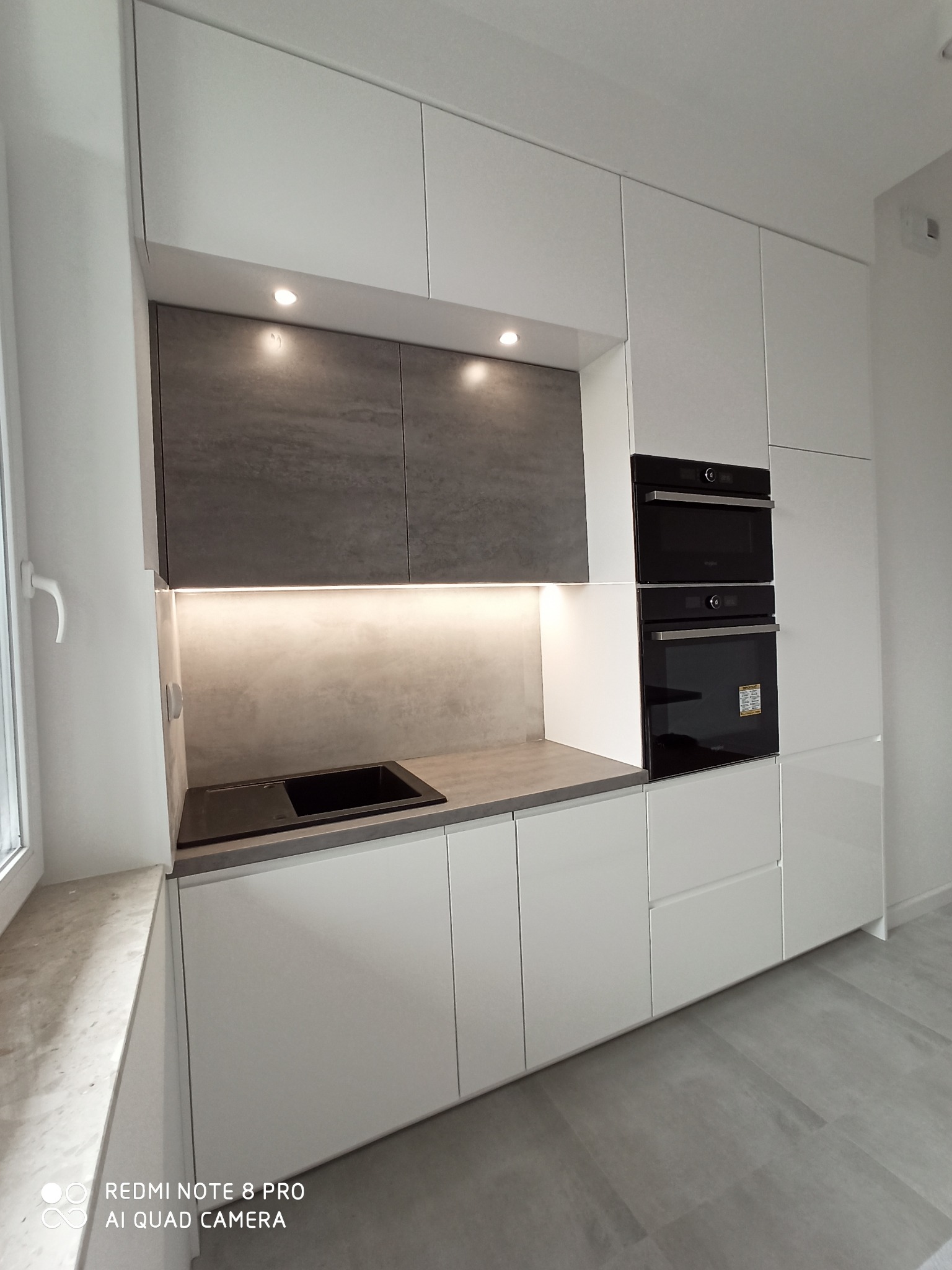 Nowoczesna kuchnia z białymi szafkami i szarym blatem, podświetlane szafki górne, zabudowane piekarniki, czarny zlew.