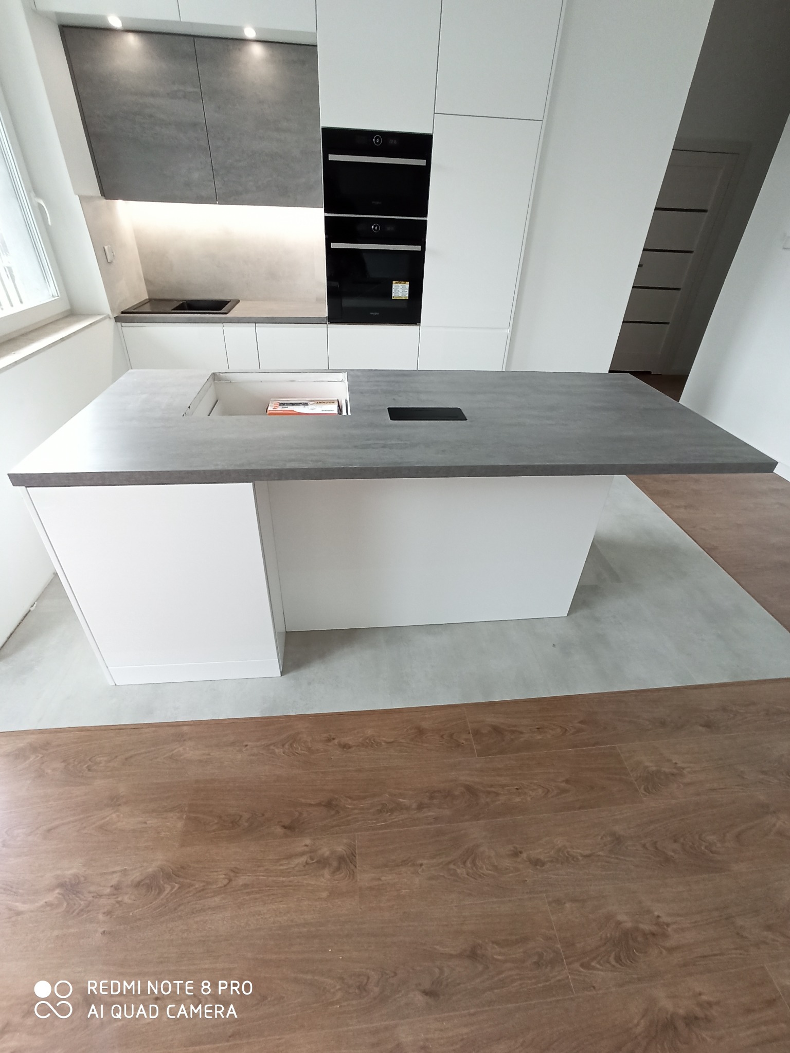 Nowoczesna kuchnia z wyspą o szarym blacie i białych frontach, widok z perspektywy podłogi z paneli imitujących drewno, w tle zabudowa kuchenna z piekarnikami.