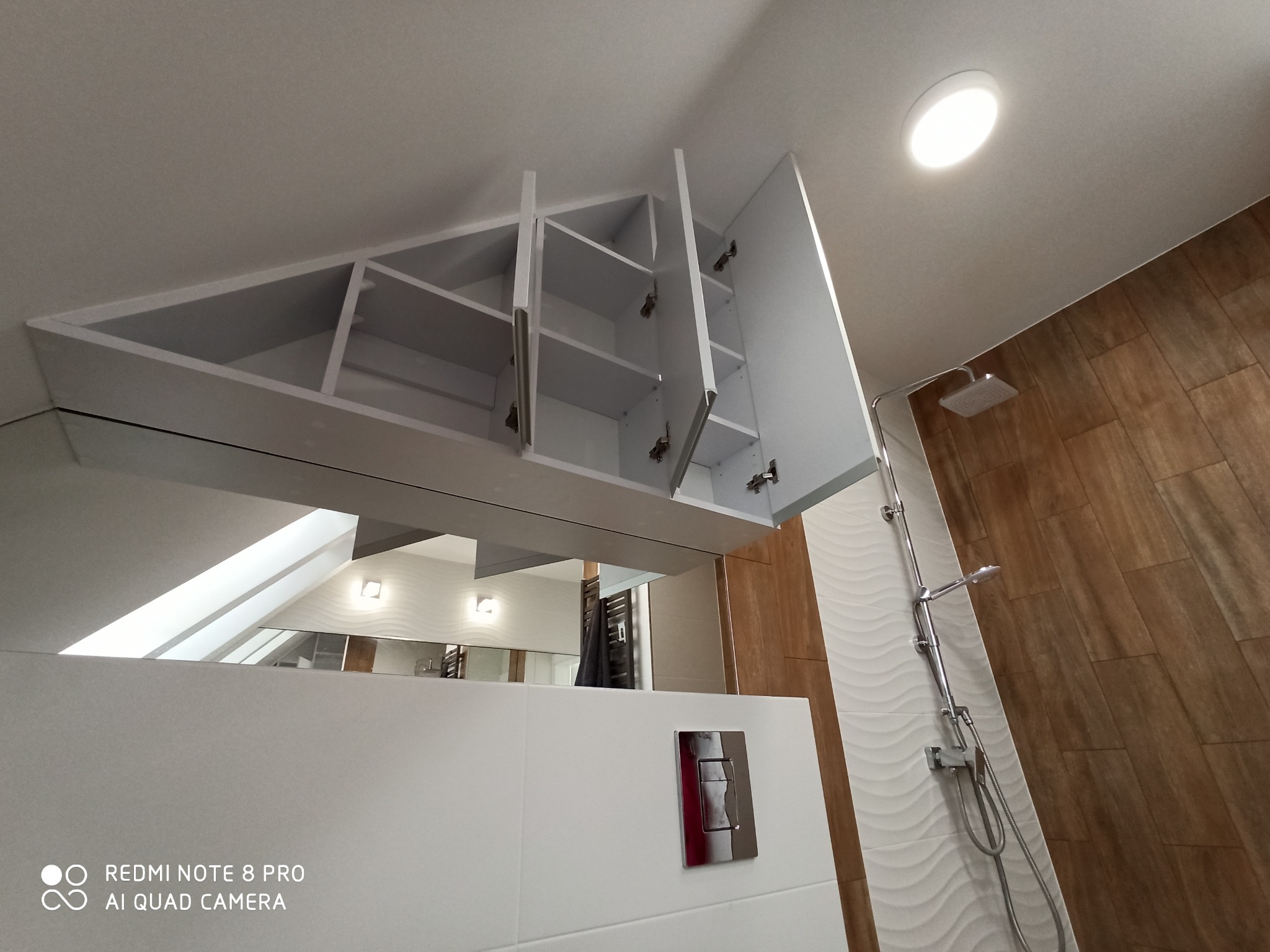 Biała szafka łazienkowa pod skosem sufitu z otwartymi drzwiczkami, widoczne półki wewnątrz, w tle kabina prysznicowa i lustro.