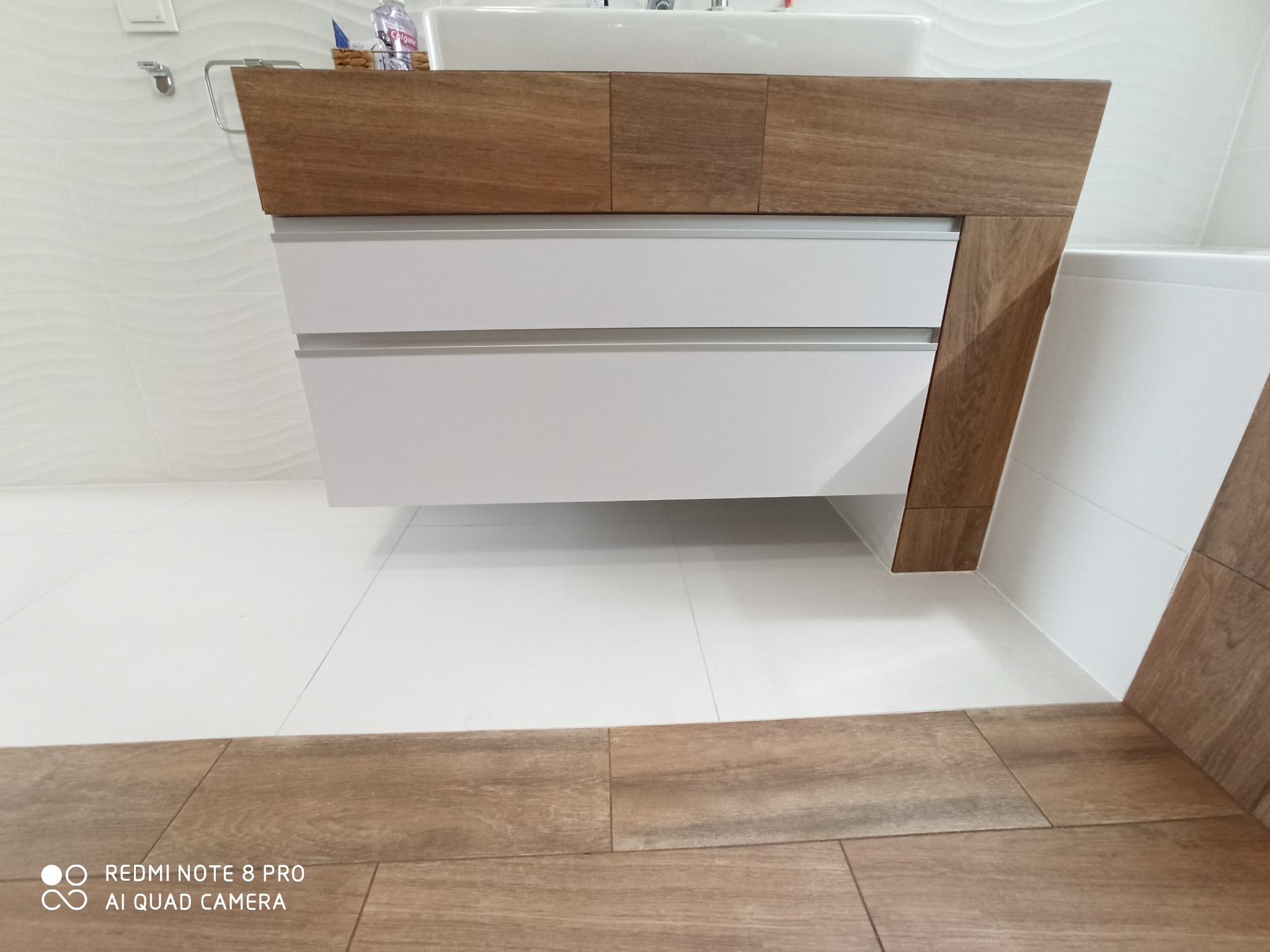 Nowoczesna szafka łazienkowa pod umywalką z trzema białymi szufladami i drewnianym blatem oraz bokiem, na tle białych płytek ściennych i podłogi z imitacji drewna.