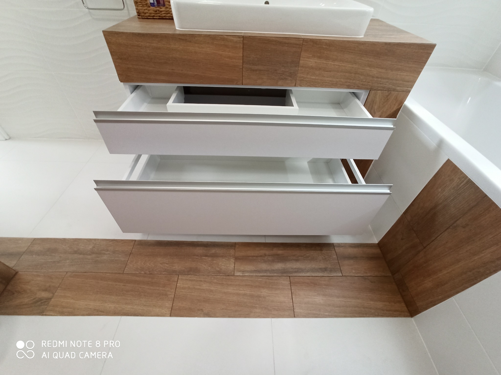 Nowoczesna szafka łazienkowa pod umywalkę z dwoma wysuwanymi szufladami o białych frontach i drewnianym korpusie, z widocznym fragmentem wanny i podłogi wyłożonej płytkami imitującymi drewno.