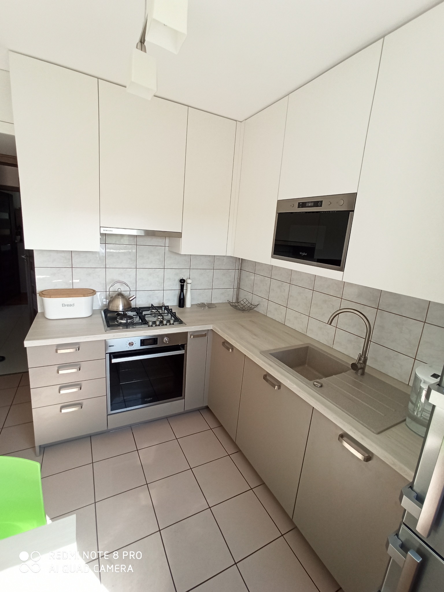 Nowoczesna kuchnia z białymi szafkami górnymi i beżowymi dolnymi, z wbudowanym piekarnikiem i mikrofalówką, oraz szarym zlewozmywakiem i baterią.