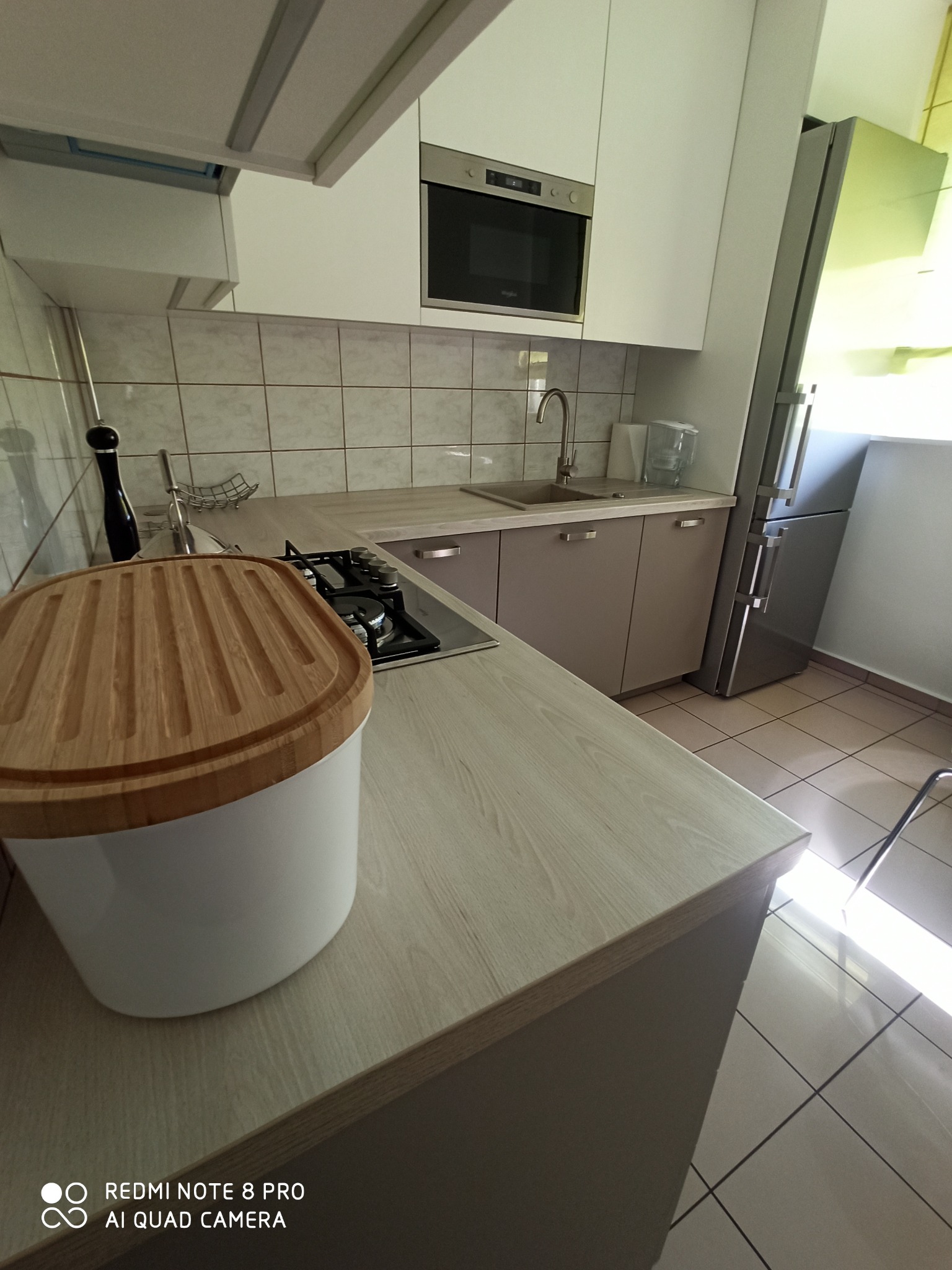 Nowoczesna kuchnia z białymi szafkami górnymi i beżowymi dolnymi, widoczny piekarnik w zabudowie, płyta gazowa, lodówka oraz pojemnik na pieczywo z drewnianą pokrywą na blacie.