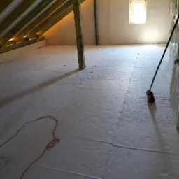 Płyty izolacyjne poddasza ułożone na podłodze, widoczna drewniana konstrukcja dachu, przewód elektryczny na podłodze i miotła oparta o ścianę.