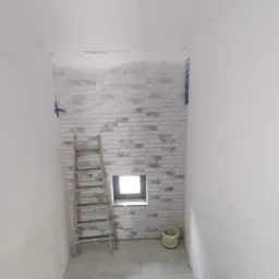 Klatka schodowa w trakcie wykończenia, ściana obłożona dekoracyjną cegłą w odcieniach bieli i szarości, widoczna drabina oparta o ścianę, wiadro z farbą i małe okno.