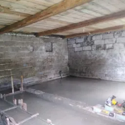 Wylewanie betonu na podłogę w piwnicy z drewnianym stropem, widoczne szalunki i narzędzia budowlane.