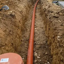 Pomarańczowa rura kanalizacyjna ułożona w wykopie ziemnym, widok z góry. W tle budynek z klimatyzatorem na ścianie.