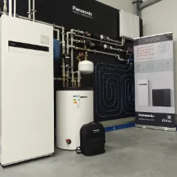 Nowoczesna instalacja grzewcza Panasonic Aquarea z pompą ciepła powietrze-woda, widoczny schemat ogrzewania podłogowego na czarnym panelu i baner reklamowy obok.