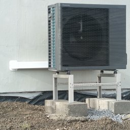 Klimatyzacja Mateusz Kurel - Zewnętrzna jednostka pompy ciepła Panasonic Aquarea zamontowana na metalowej konstrukcji, na betonowych bloczkach, przy ścianie budynku. Widoczny biały przewód.