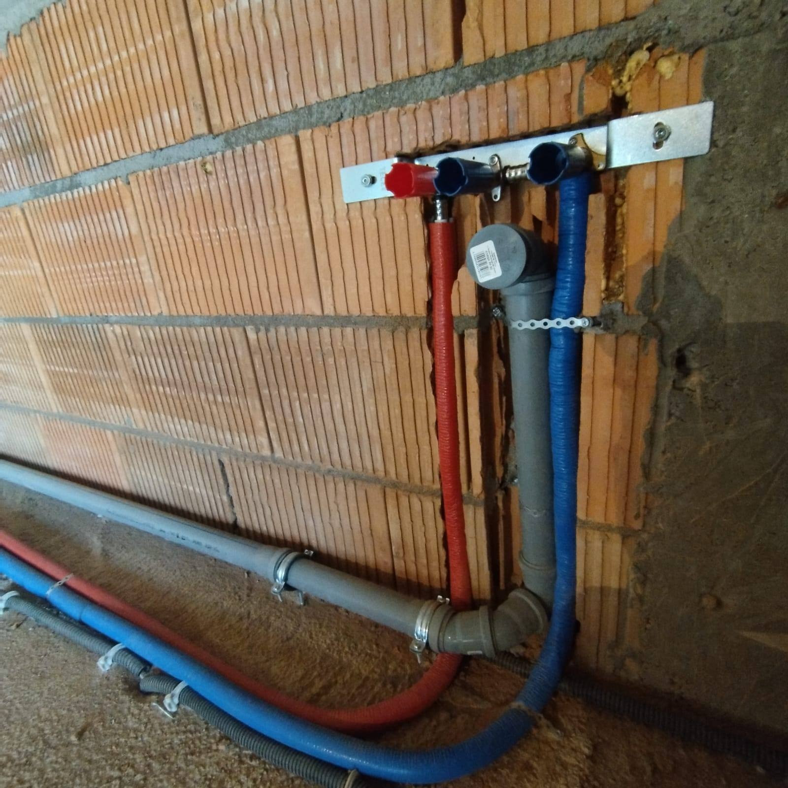Fragment instalacji wodnej z rurami PEX w kolorach czerwonym i niebieskim, zamocowanymi do ściany z cegły dziurawki za pomocą metalowej szyny, widoczne również szare rury kanalizacyjne