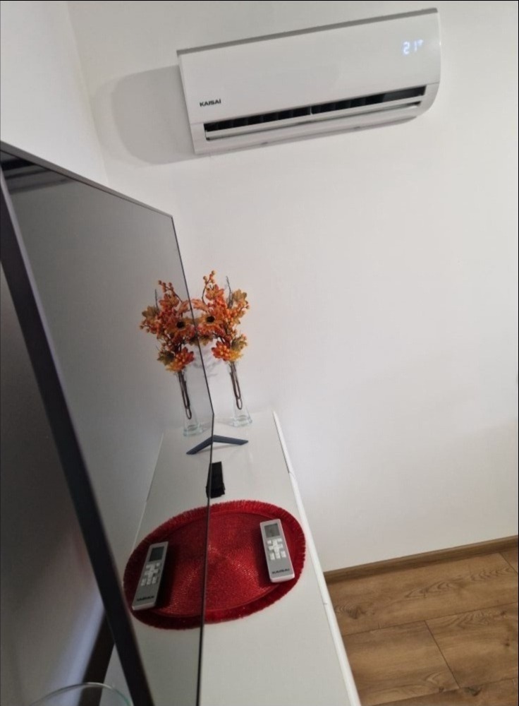 Biały klimatyzator marki Kaisai zamontowany na ścianie nad białą komodą z dekoracjami, w tle widoczna podłoga z paneli.