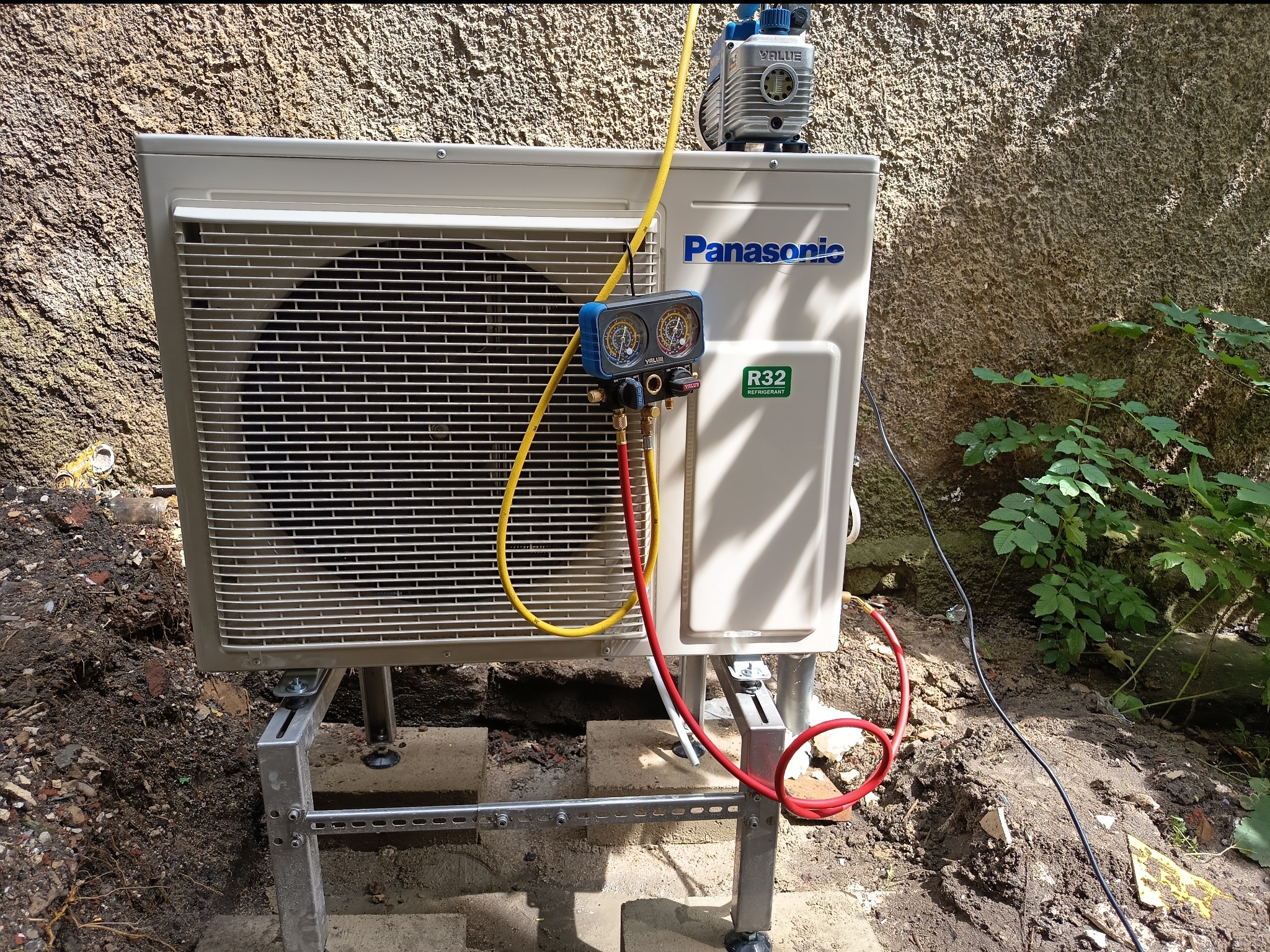 Biała jednostka zewnętrzna klimatyzacji Panasonic, zamontowana na metalowym stelażu i podłączona do manometru oraz pompy próżniowej R32 na tle ściany i zieleni.