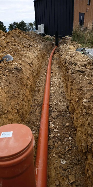 Pomarańczowa rura kanalizacyjna ułożona w wykopie ziemnym, widok z góry. W tle budynek z klimatyzatorem na ścianie.