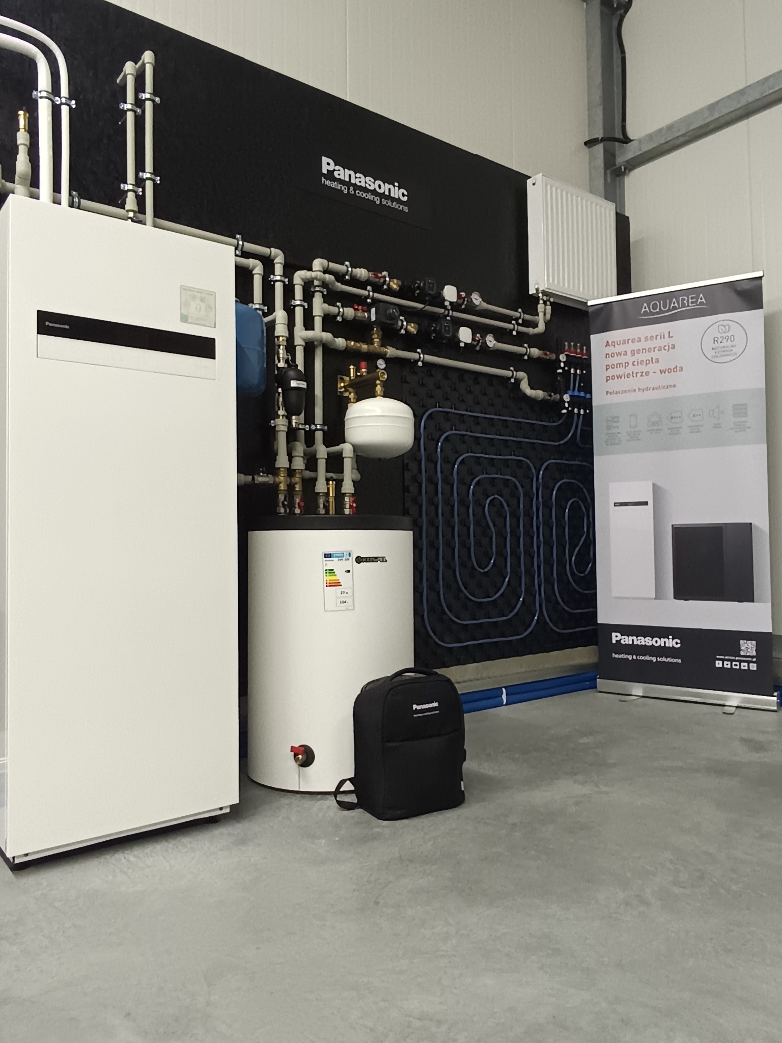 Nowoczesna instalacja grzewcza Panasonic Aquarea z pompą ciepła powietrze-woda, widoczny schemat ogrzewania podłogowego na czarnym panelu i baner reklamowy obok.