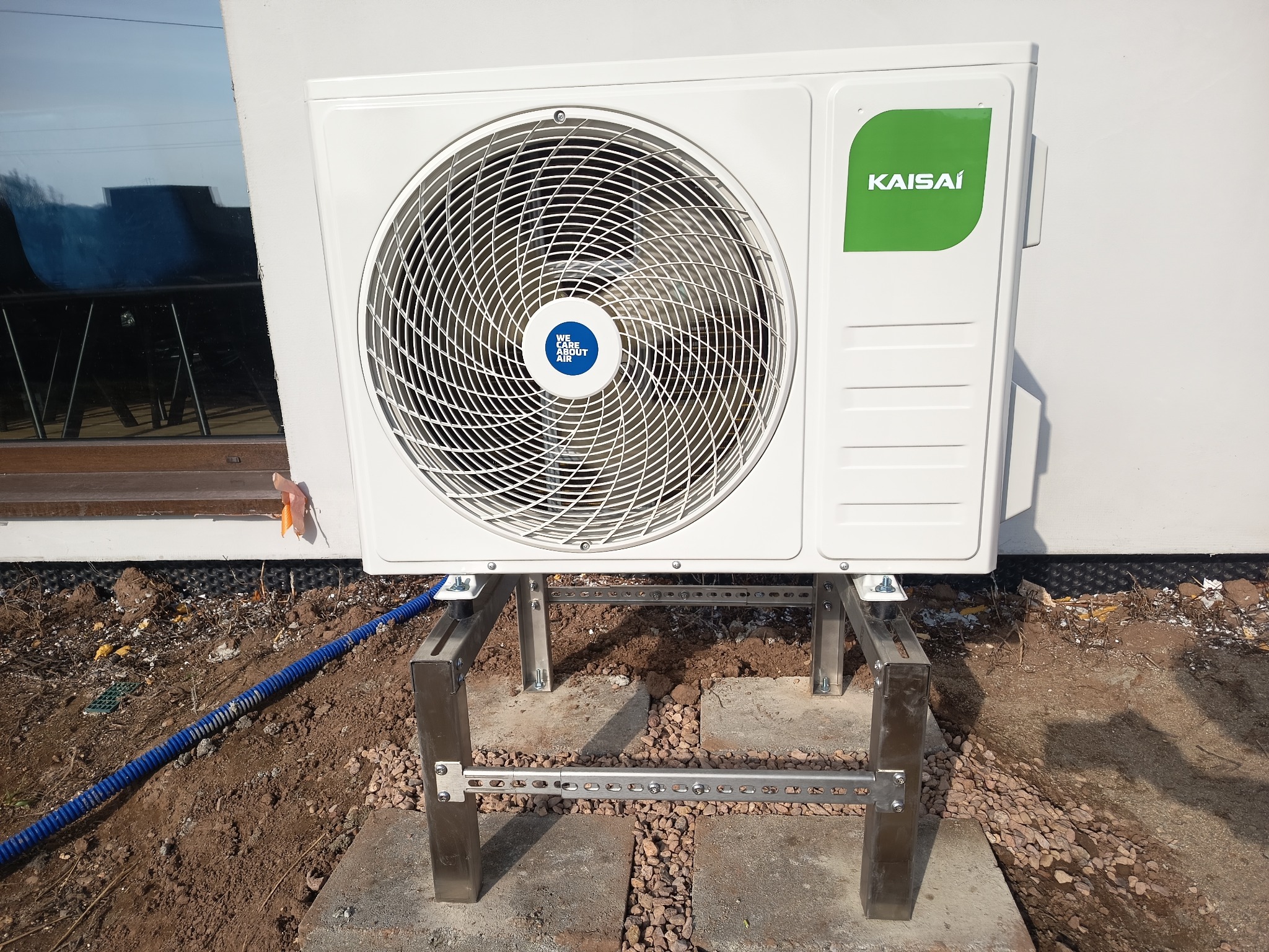 Biała jednostka zewnętrzna klimatyzacji Kaisai, zamontowana na metalowym stelażu, z widocznym niebieskim przewodem i fragmentem okna w tle. Ujęcie z poziomu gruntu.