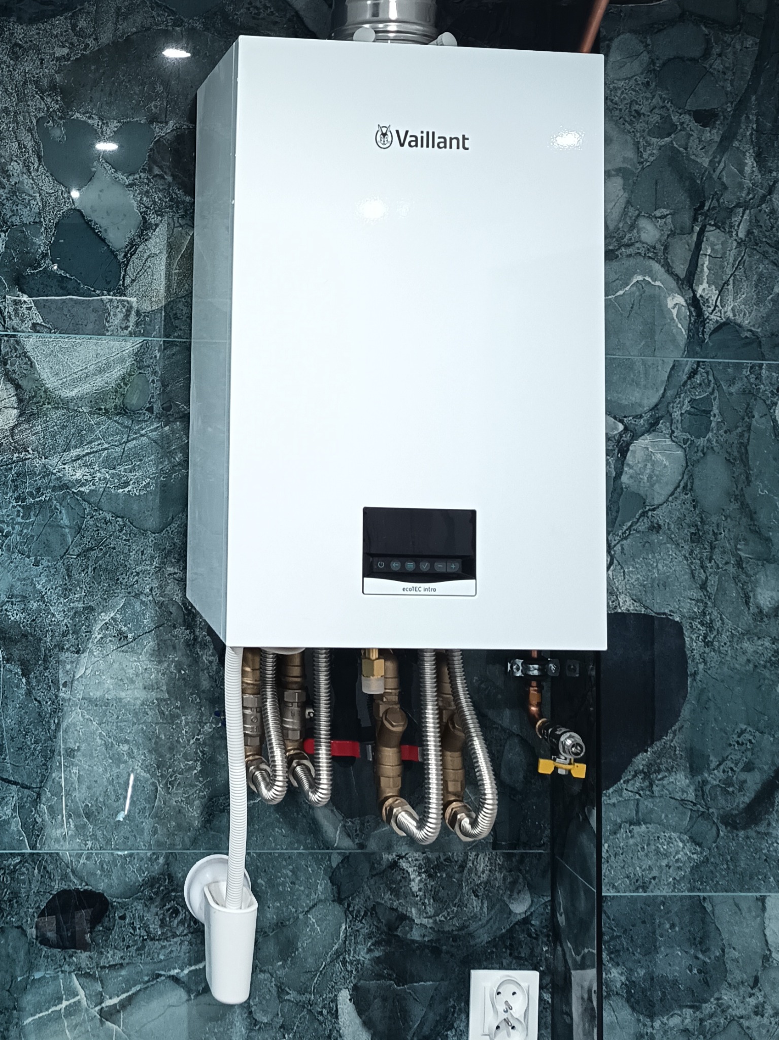 Biały, wiszący piec gazowy Vaillant z podłączonymi rurami na tle ściany z płytek imitujących marmur. Widoczny fragment instalacji i gniazdko elektryczne.
