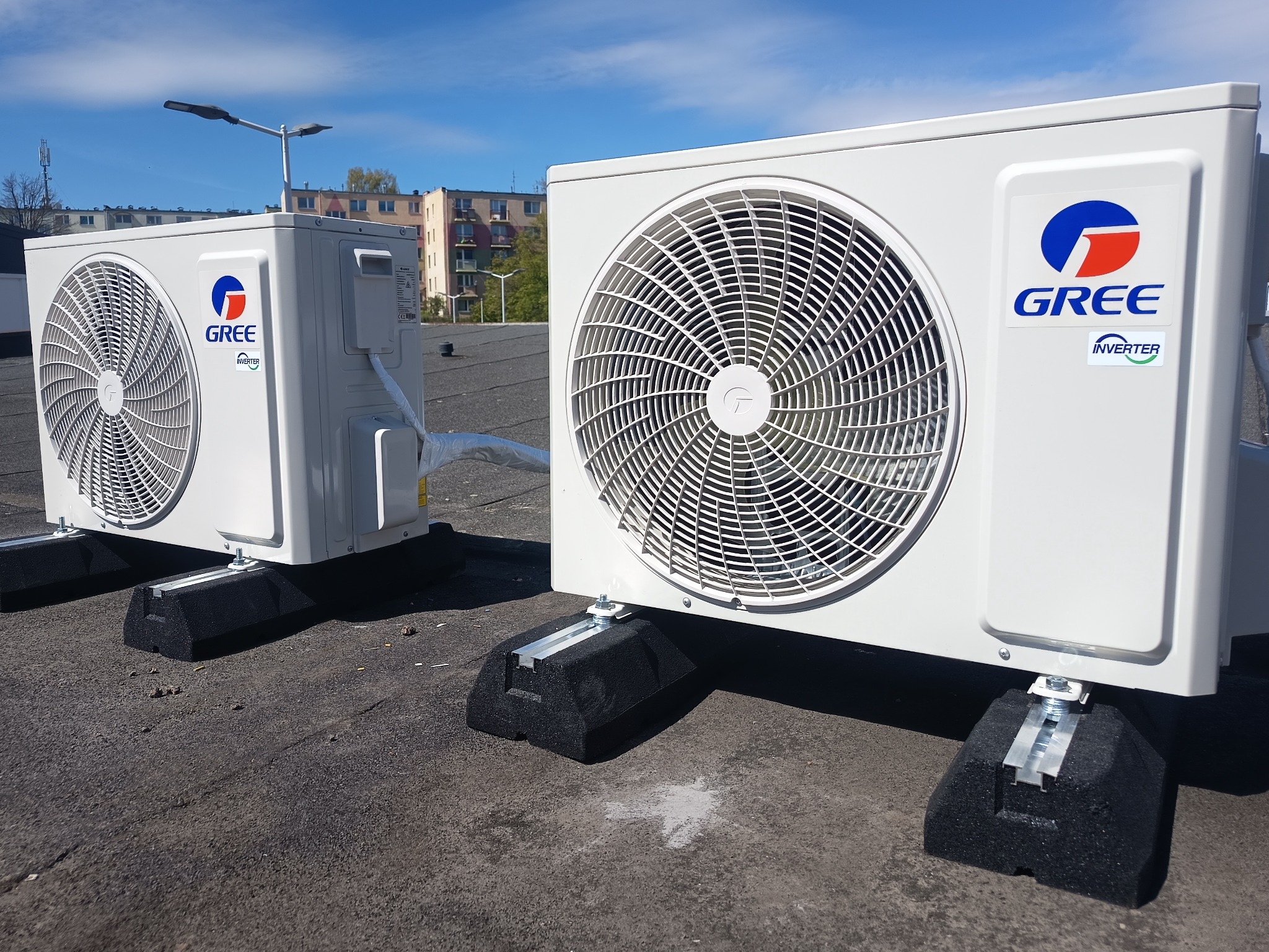 Dwa białe klimatyzatory Gree Inverter na dachu budynku, zamontowane na czarnych, gumowych podstawach. Błękitne niebo w tle. Widoczne detale montażu.