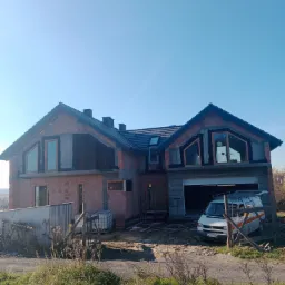 Surowy stan domu murowanego z garażem wbudowanym, widoczne okna z pomarańczowymi ramami i czarna dachówka, zaparkowany biały bus przed garażem, widok z poziomu gruntu na tle błękitnego nieba.