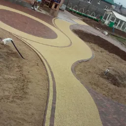 Kręty chodnik z jasnożółtej kostki brukowej obramowany brązową kostką, prowadzący od drewnianej altany w kierunku szklarni w pochmurny dzień.