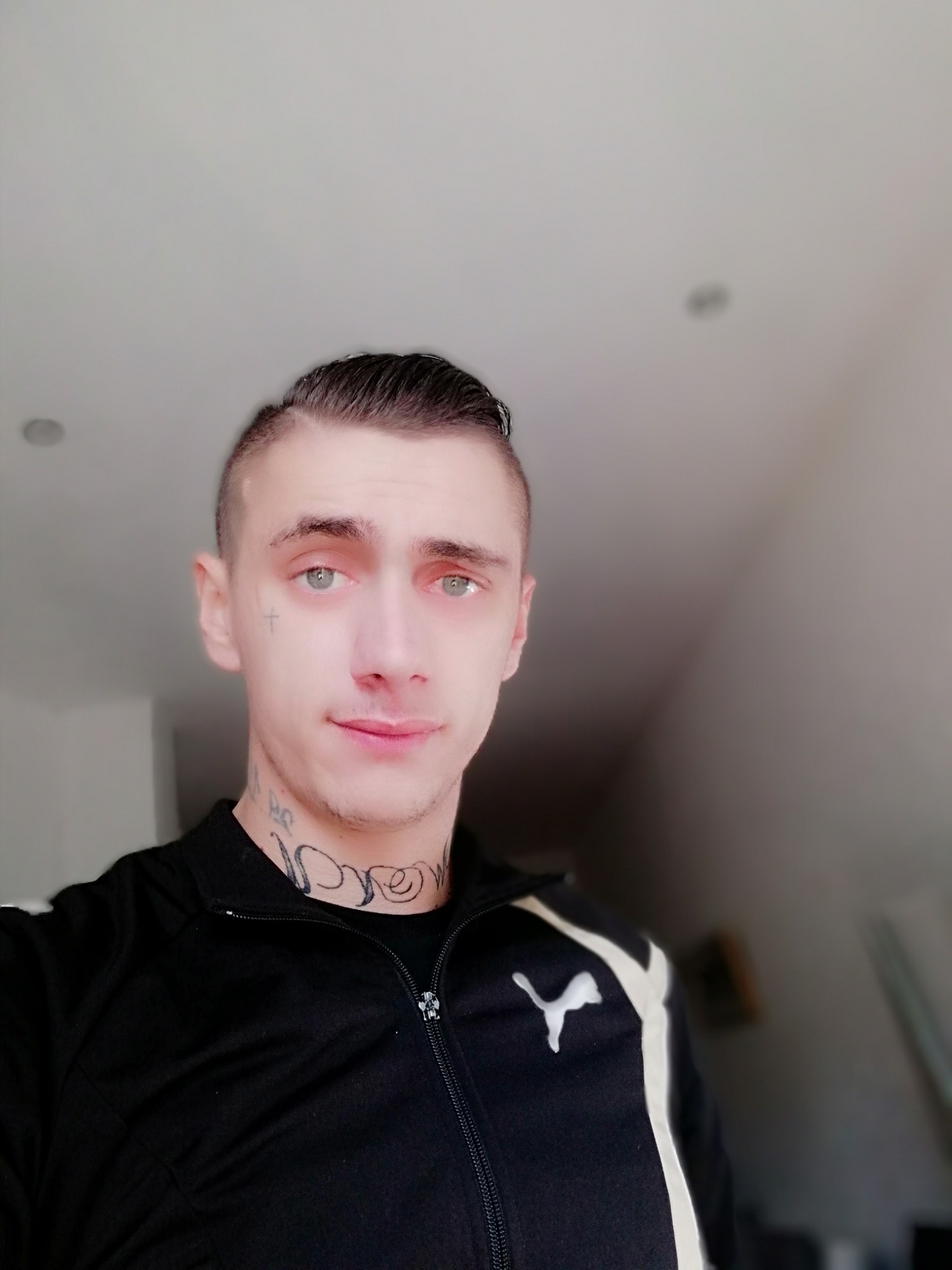 Portret młodego mężczyzny z tatuażami na twarzy i szyi, ubranego w czarną bluzę sportową Puma w Hamm, z widocznym krzyżykiem wytatuowanym pod okiem.