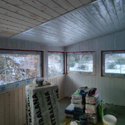 Wnętrze pomieszczenia w trakcie ocieplania poddasza, widoczne płyty izolacyjne, worki z zaprawą, drewniana boazeria na ścianach i suficie oraz okna z widokiem na zimowy krajobraz.