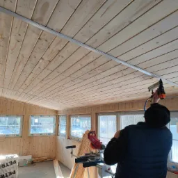 Wnętrze wykończone drewnianą boazerią, pracownik mocuje listwę podsufitową za pomocą narzędzia pneumatycznego, widoczne okna z widokiem na zimowy krajobraz, na podłodze materiały budowlane i piła...