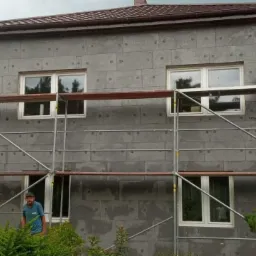 Dom w trakcie ocieplania szarą wełną mineralną, widoczne rusztowania, pracownik w niebieskiej koszulce stoi przy krzewach na pierwszym planie.