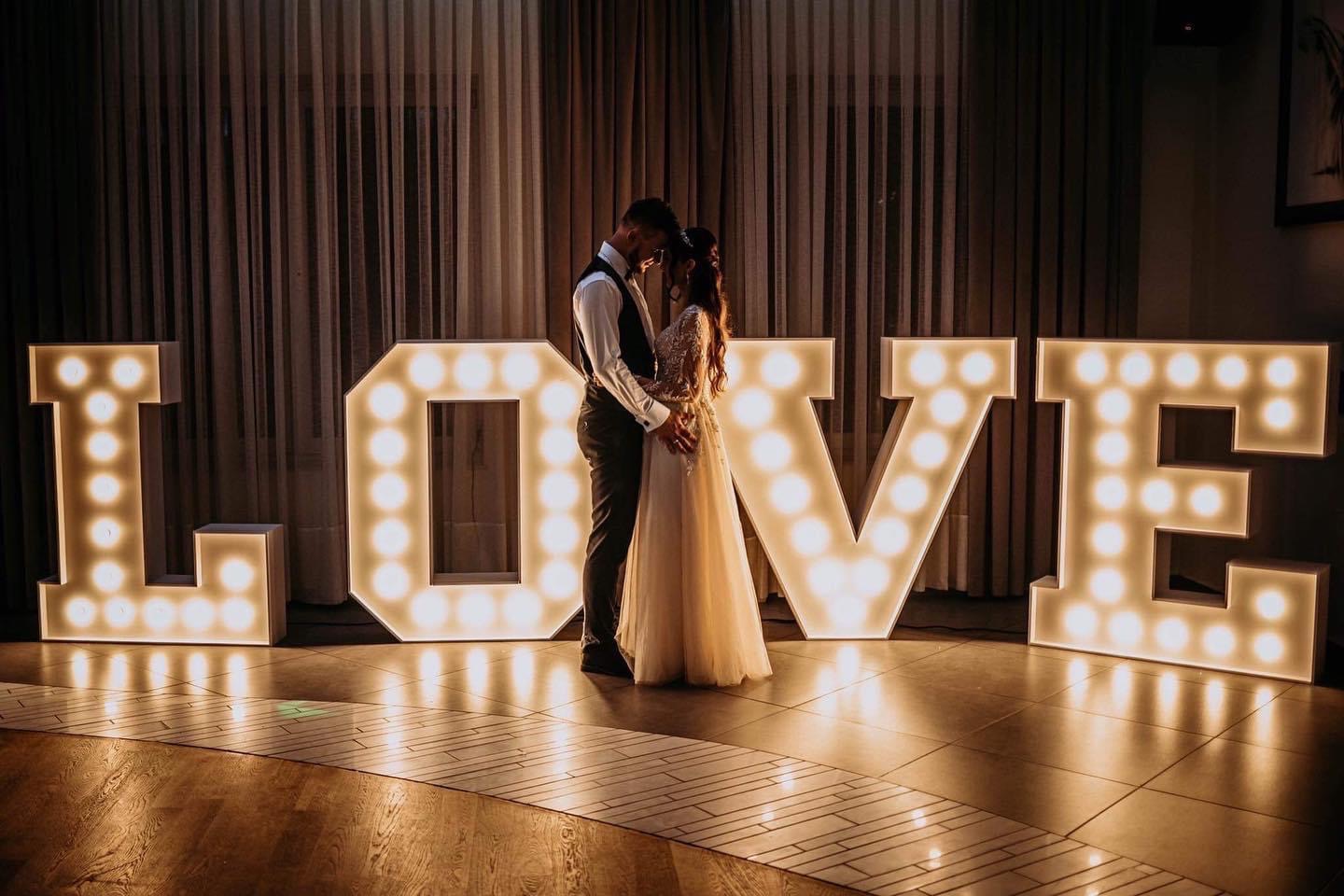 Para młoda obejmująca się przed podświetlanym napisem LOVE z żarówkami, ustawionym na parkiecie z wzorem w jodełkę, na tle zasłon.