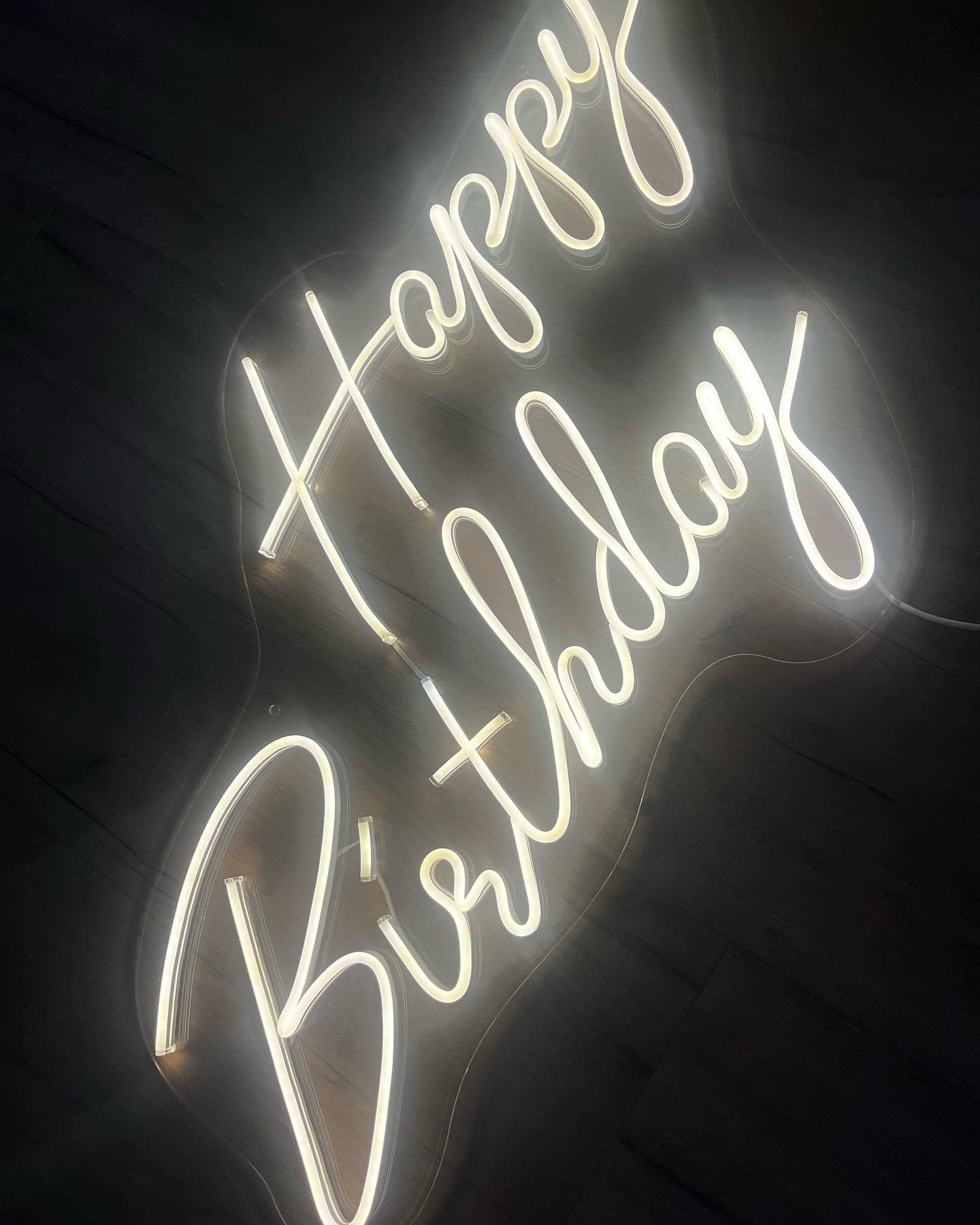 Neonowy napis 'Happy Birthday' na ciemnym, drewnianym tle, delikatnie oświetlony, z widocznym kablem zasilającym po prawej stronie.