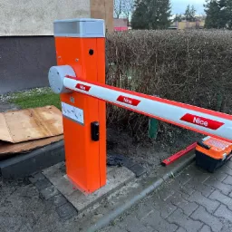 Pomarańczowa bariera automatyczna do bramy z napisem 'NICE', zamontowana na betonowej podstawie, obok leżą narzędzia i poziomica, widoczne ślady po deszczu.