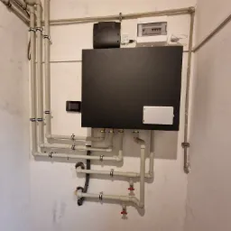 Czarna jednostka centralna z rurami i zaworami w piwnicy, widoczna skrzynka elektryczna i rury wzdłuż ściany, szare ściany z widocznymi śladami wilgoci.