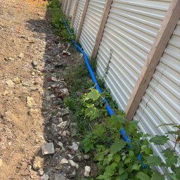 Fotowoltaika Golta Sp.z o.o - Niebieski przewód w ziemi wzdłuż ogrodzenia z blachy trapezowej, prawdopodobnie instalacja elektryczna zewnętrzna, widoczny pracownik w oddali.