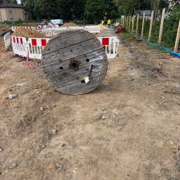 Fotowoltaika Golta Sp.z o.o - Duża, drewniana szpula na kable elektryczne leży na ziemi na placu budowy. W tle widoczne barierki, budynek i pracownicy w kamizelkach odblaskowych.