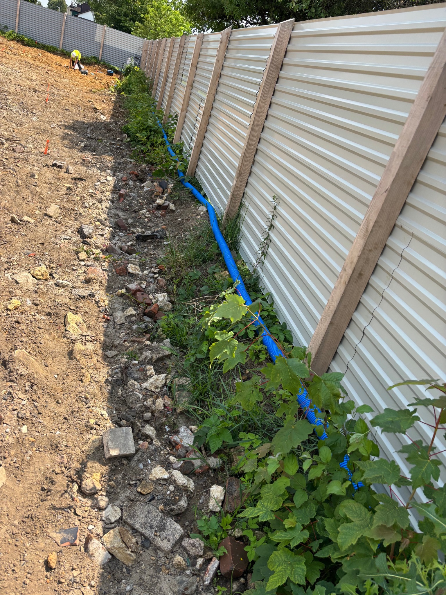 Niebieski przewód w ziemi wzdłuż ogrodzenia z blachy trapezowej, prawdopodobnie instalacja elektryczna zewnętrzna, widoczny pracownik w oddali.