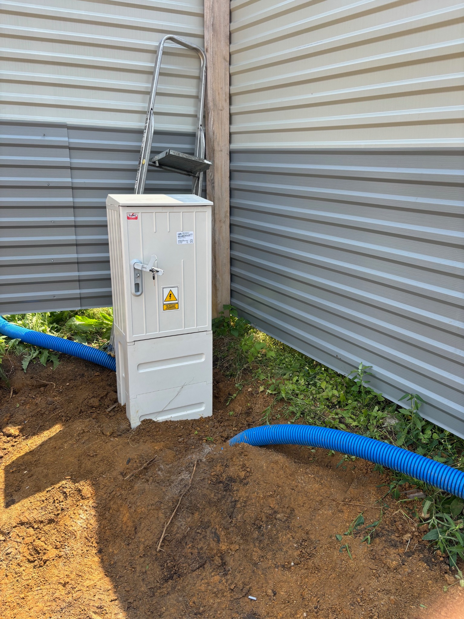 Biała skrzynka elektryczna z ostrzeżeniem, zainstalowana na ziemi obok metalowego ogrodzenia. Widoczne niebieskie rury ochronne i drabina oparta o słupek.