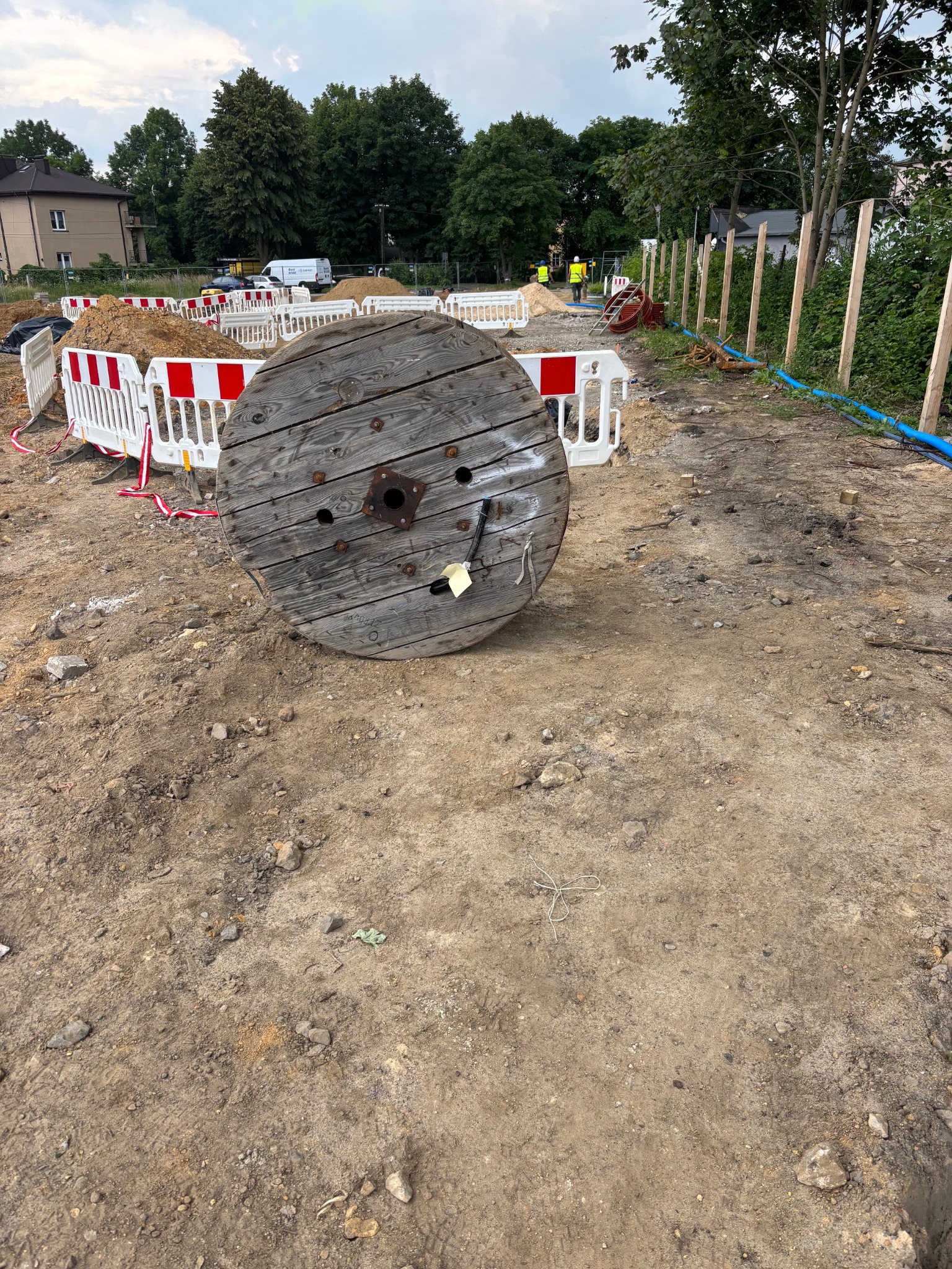 Duża, drewniana szpula na kable elektryczne leży na ziemi na placu budowy. W tle widoczne barierki, budynek i pracownicy w kamizelkach odblaskowych.