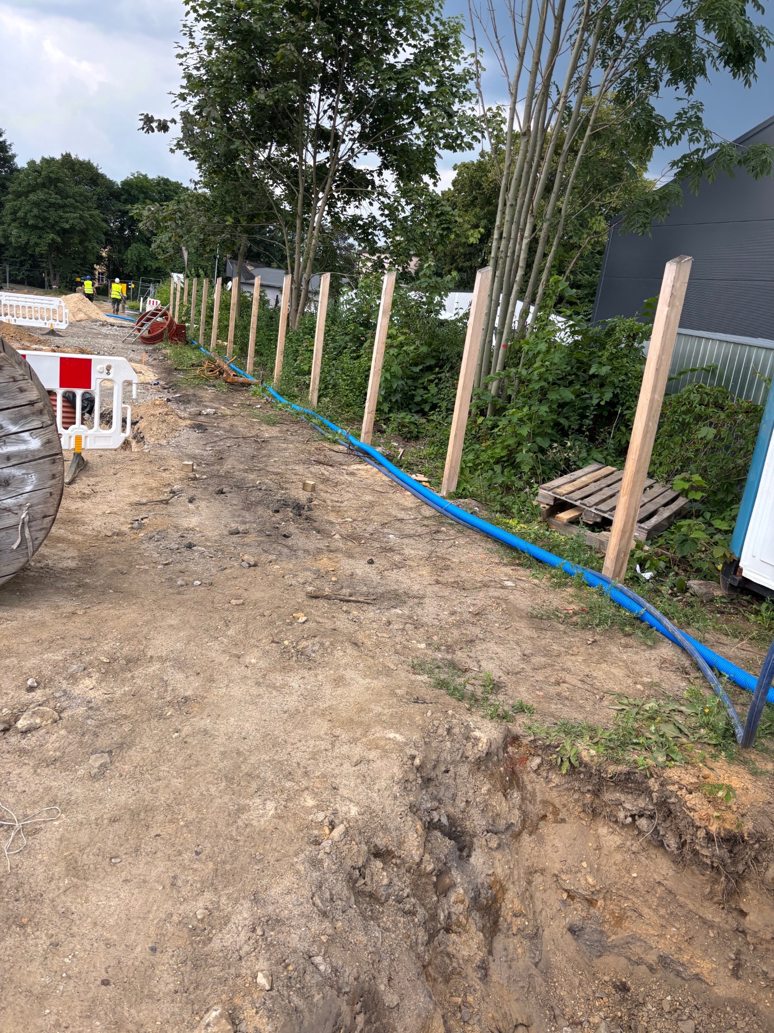 Niebieski przewód ułożony wzdłuż wykopu, zabezpieczony drewnianymi słupkami. W tle widoczna zieleń i budynek. Prace ziemne w toku.