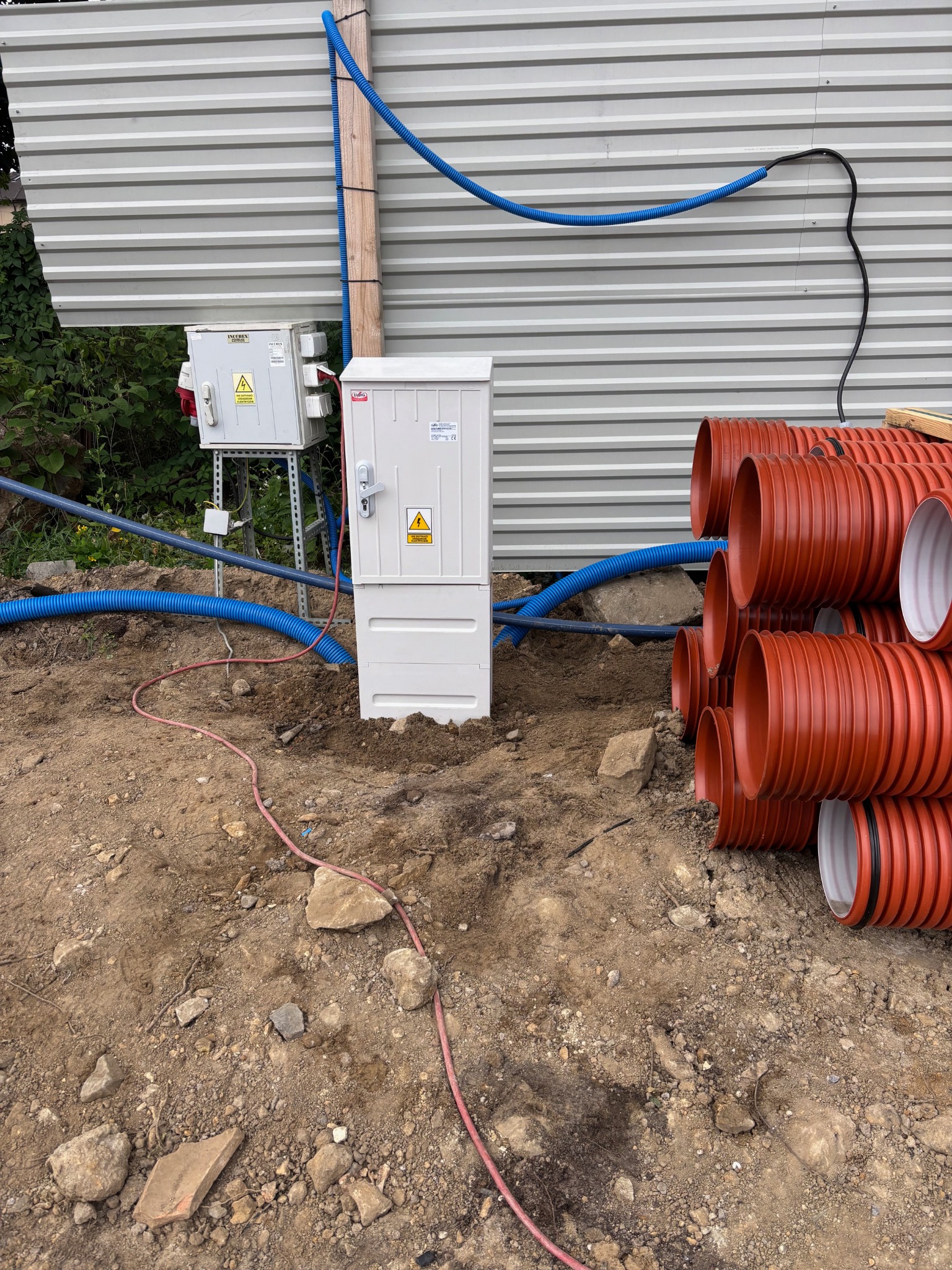 Biała skrzynka elektryczna z ostrzeżeniem o napięciu, obok pomarańczowe rury karbowane i szara blacha falista w tle. Widoczne kable i przewody.