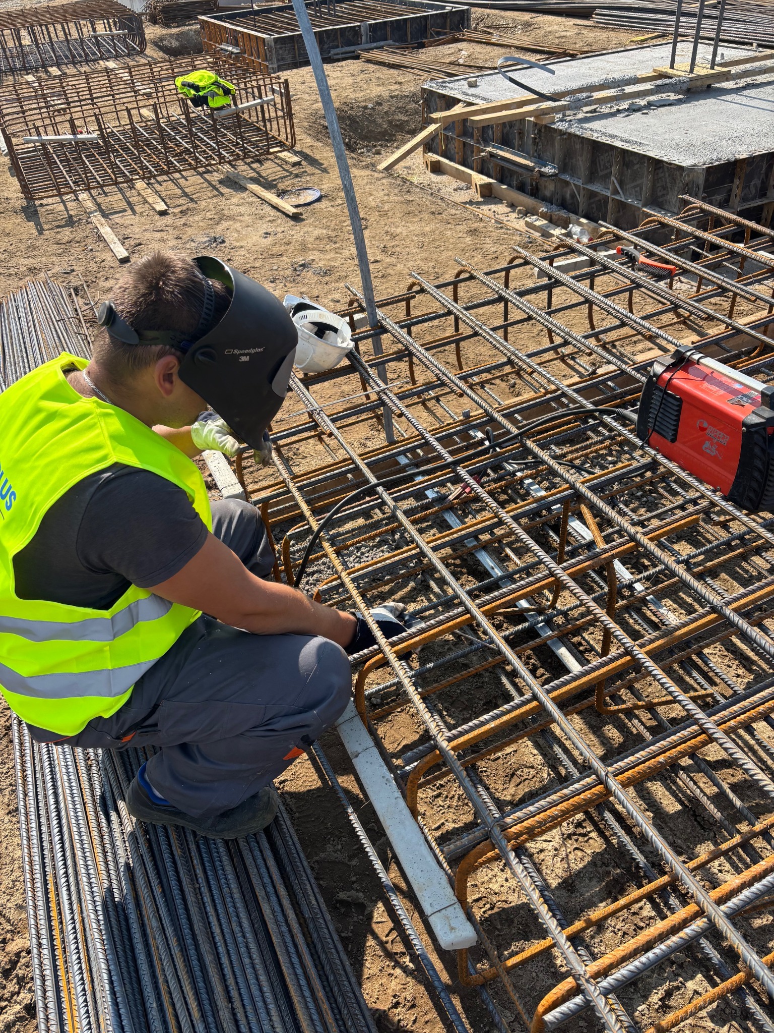 Spawanie bednarki do zbrojenia pod instalację odgromową. Widoczny spawacz w masce i kamizelce, zbrojenie, spawarka i pręty zbrojeniowe na ziemi.