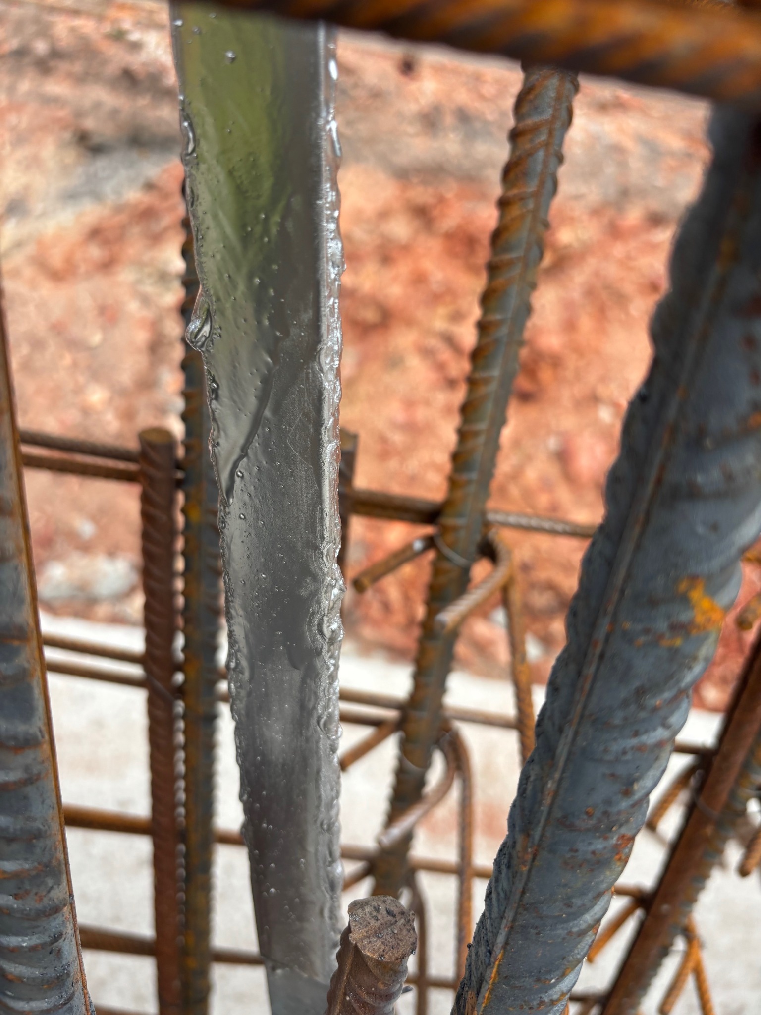 Z bliska: stalowy płaskownik z kroplami wody, część konstrukcji odgromowej na tle zbrojenia i gruntu. Widok z góry, ostrość na pierwszym planie.