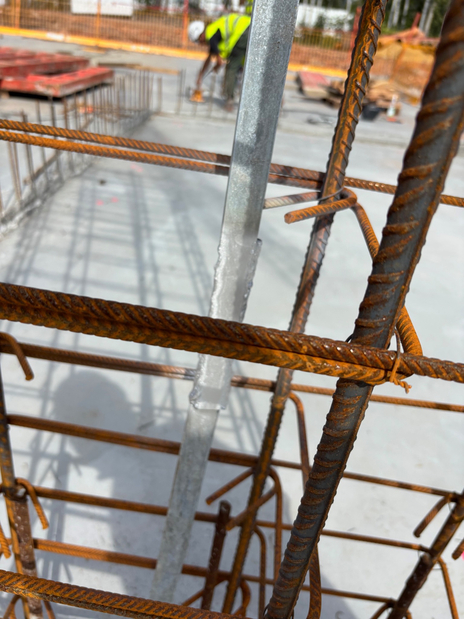 Zbrojenie fundamentów z widocznymi prętami zbrojeniowymi i elementami konstrukcyjnymi na budowie, z pracownikiem w tle. Widok z bliska na siatkę zbrojeniową.