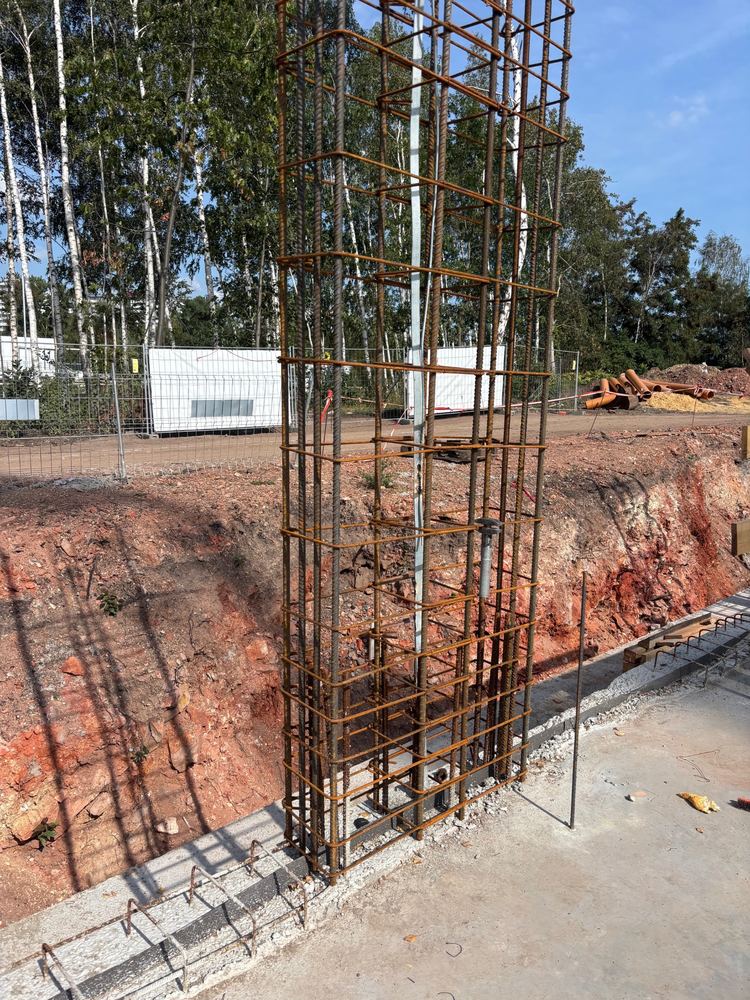 Zbrojenie kolumny na fundamencie, widok z góry. Konstrukcja z prętów stalowych na tle wykopu i drzew. Prace przygotowawcze do wylania betonu.