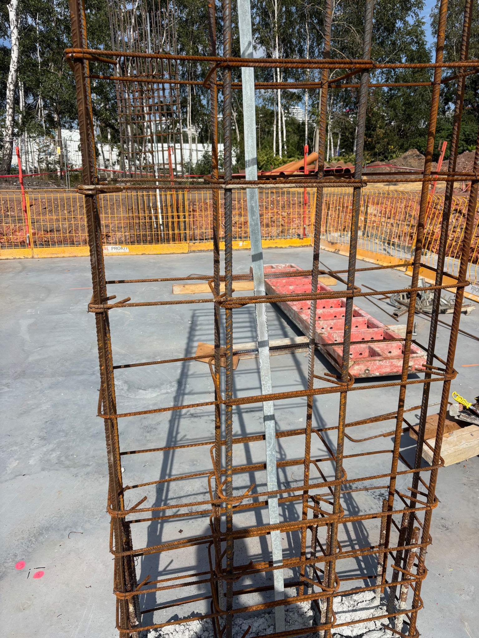 Zbrojenie kolumny na betonowym fundamencie, zabezpieczone metalowym profilem. W tle widoczne szalunki i drzewa.