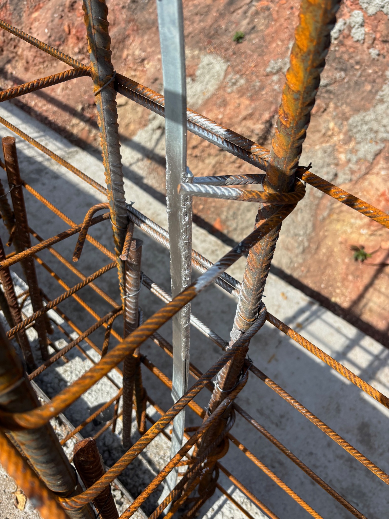 Z bliska: Zbrojenie fundamentów z prętów żebrowanych połączonych drutem, z elementem metalowym. Widoczne ślady korozji na prętach i beton w tle.